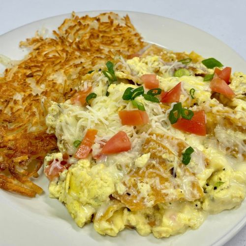 A plate of scrambled eggs with cheese, tomatoes, and green onions, served with crispy hash browns on the side.