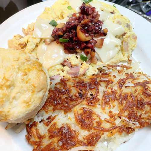 A plate of scrambled eggs with cheese, topped with diced fruit, served with a biscuit and crispy hash browns.