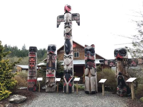A collection of tall totem poles in a natural setting, with a building and trees in the background.