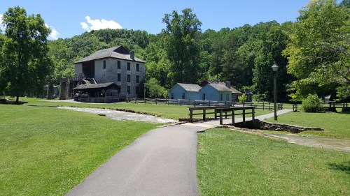 A scenic view of a historic building and blue house surrounded by greenery, with a path and stream in the foreground.