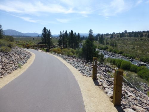 A winding path alongside a river, surrounded by trees and mountains under a clear blue sky.