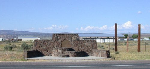 A stone monument in an open field, with a plaque, surrounded by grass and distant buildings under a blue sky.