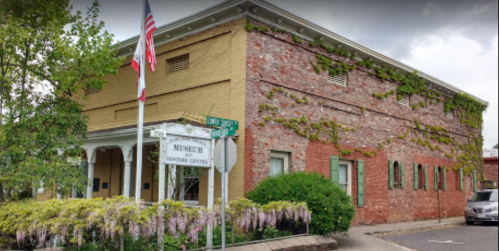 Historic brick building with green shutters, ivy-covered walls, and a sign for a museum, surrounded by blooming flowers.