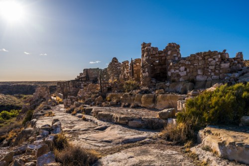 Ruins of an ancient stone structure under a bright sun, surrounded by rocky terrain and sparse vegetation.