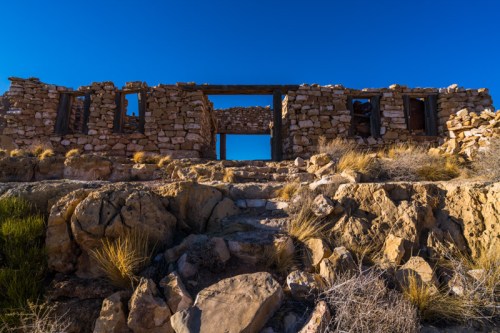 Ruins of a stone building on rocky terrain, framed by a clear blue sky.