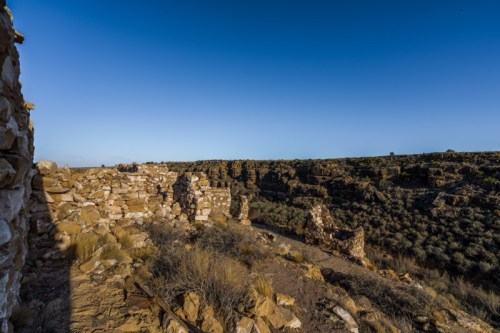 Ruins of a stone structure on a rocky landscape under a clear blue sky, surrounded by sparse vegetation.