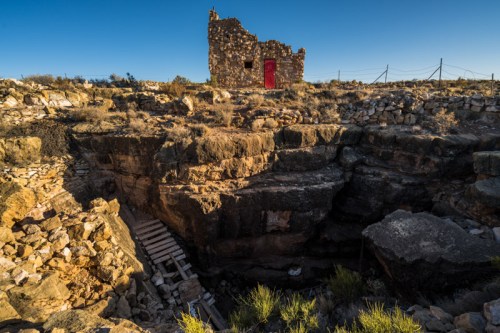 A rocky landscape features a ruined stone building with a red door, overlooking a deep, rugged pit.