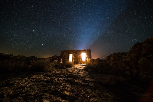 A starry night sky above a dilapidated stone building with glowing windows, surrounded by rocky terrain.