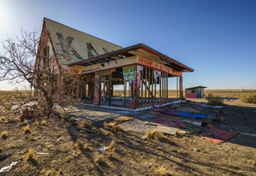 Abandoned building with graffiti, surrounded by dry land and debris under a clear blue sky.