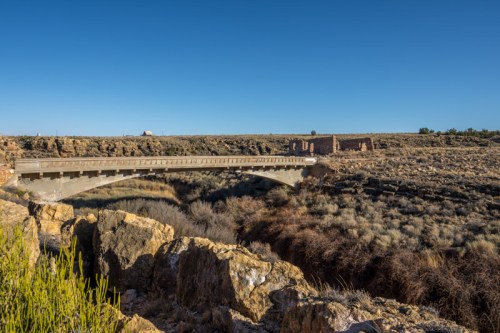 A bridge spans a dry canyon, with rocky terrain and sparse vegetation under a clear blue sky.