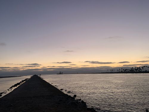 A serene view of a pier extending into calm waters at sunset, with clouds and palm trees in the background.