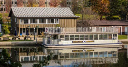A boat named "Scenic City Empress" docked by a riverside building, reflecting in calm water. Trees and buildings in the background.