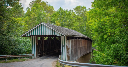 A rustic covered bridge surrounded by lush green trees and a winding road.