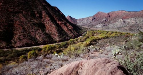A scenic view of red rock mountains and a winding river surrounded by green vegetation under a clear blue sky.