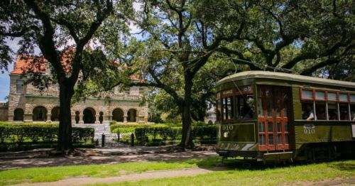 A vintage streetcar passes by a historic building surrounded by lush trees and greenery on a sunny day.