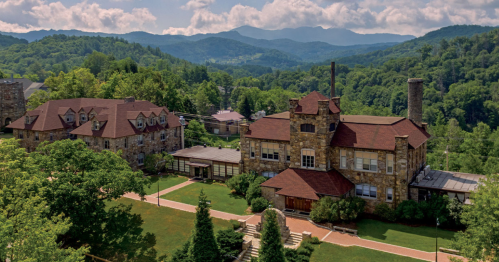 Aerial view of a stone building surrounded by lush greenery and mountains under a partly cloudy sky.