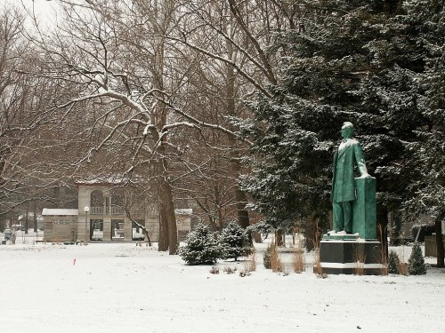 A snow-covered park scene featuring a statue and trees, with a building visible in the background.