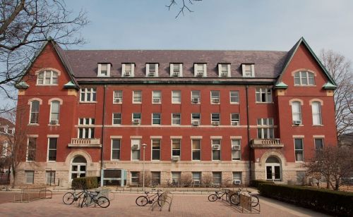 A large red brick building with multiple windows and a peaked roof, surrounded by trees and bicycles parked outside.