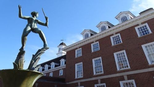 A bronze statue of a figure holding a stick stands in front of a brick building under a clear blue sky.
