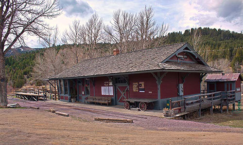 Historic red train station surrounded by trees and mountains, with a wooden walkway and wagon in front.