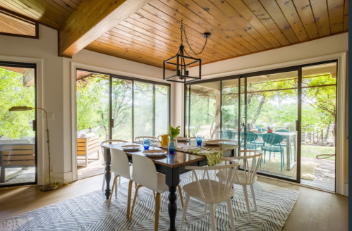 Bright dining area with a wooden table, white chairs, and large windows overlooking a green outdoor space.