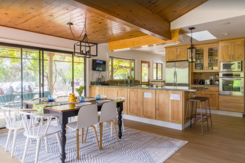Bright kitchen with wooden accents, a dining table, and large windows overlooking a garden. Modern appliances included.