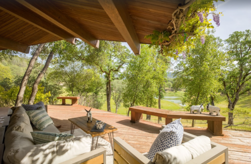 A cozy outdoor seating area with wooden furniture, surrounded by lush greenery and a scenic view of a pond.