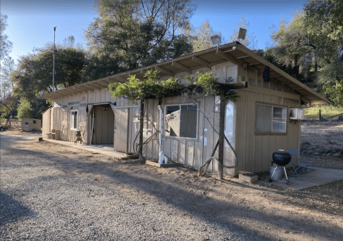 A rustic, single-story building with a gravel path, surrounded by trees and outdoor equipment.