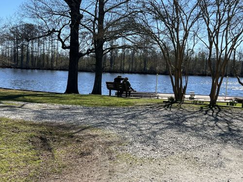 A couple sits on a bench by a calm river, surrounded by trees and a clear blue sky.