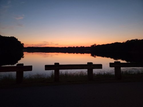 A serene lake at sunset, reflecting vibrant colors in the water, with a wooden barrier in the foreground.