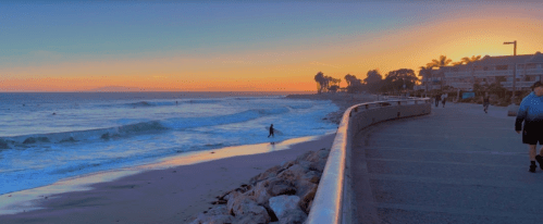 A serene beach at sunset, with gentle waves, a surfer, and a walkway lined with palm trees and people.