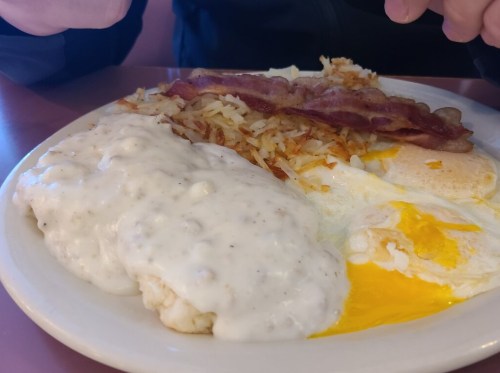 A plate of breakfast featuring biscuits with gravy, fried eggs, crispy bacon, and hash browns.
