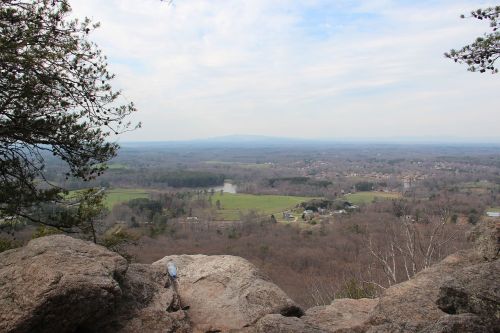 A scenic view from a rocky overlook, showcasing rolling hills, a river, and a distant mountain range under a cloudy sky.