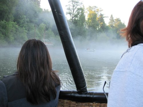 Two people view a misty river scene from a boat, with trees and a distant boat visible in the background.