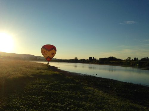 A colorful hot air balloon floats above a calm river at sunrise, with lush greenery along the shore.