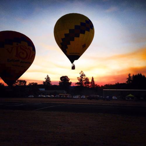 Two hot air balloons silhouetted against a colorful sunset, with trees and parked cars in the background.