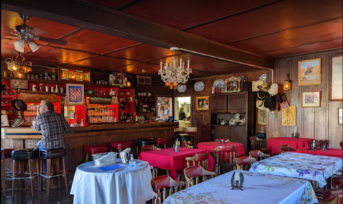 A rustic bar interior with red tablecloths, a chandelier, and a lone patron sitting at the counter.