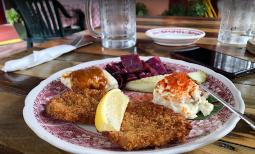 A plate of fried fish with lemon, coleslaw, pickles, and purple cabbage on a wooden table.