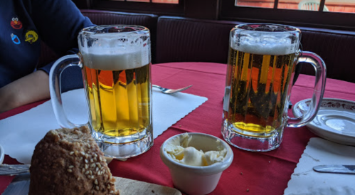 Two frosty mugs of beer on a red tablecloth, accompanied by a roll and a small dish of butter.