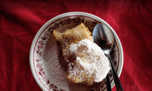 A slice of dessert topped with whipped cream and powdered sugar, served on a decorative plate with a spoon.