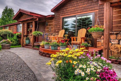 A charming wooden house with a flower-filled garden, featuring two Adirondack chairs on a welcoming porch.