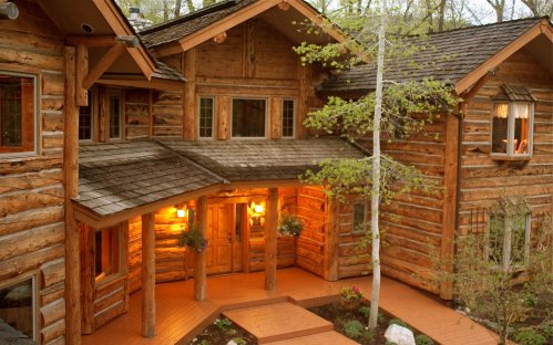 A rustic log cabin with a welcoming entrance, surrounded by trees and landscaped gardens. Warm lights glow from the windows.