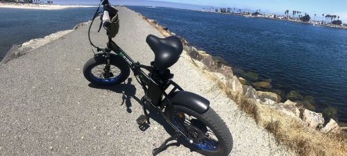 A bike rests on a rocky path beside a calm waterway, with palm trees and a distant shoreline in the background.