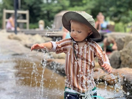 A young child in a hat plays in a splash pad, reaching out to touch the water jets.