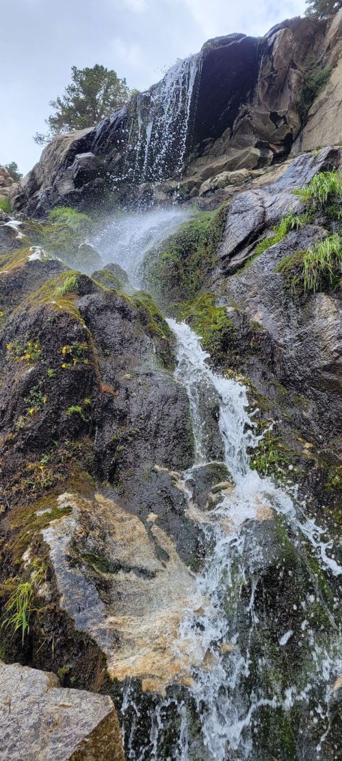 A cascading waterfall flows over rocky terrain, surrounded by lush greenery and mist in a natural setting.
