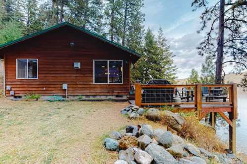 A wooden cabin by a river, surrounded by trees, with a deck and rocky shoreline.