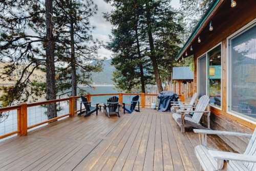 A wooden deck with Adirondack chairs overlooks a serene lake surrounded by trees and mountains.