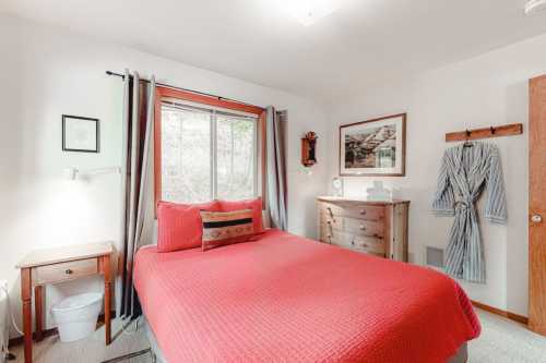 Cozy bedroom with a red bedspread, window, dresser, and robe hanging on the wall. Natural light fills the space.