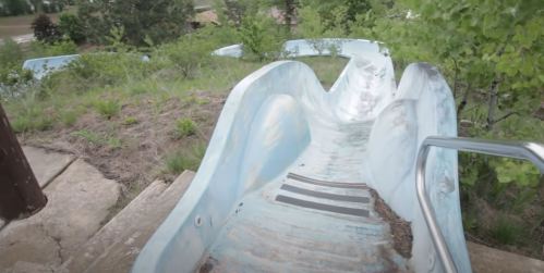 An abandoned water slide winding through overgrown greenery, with a weathered blue surface and metal railing.