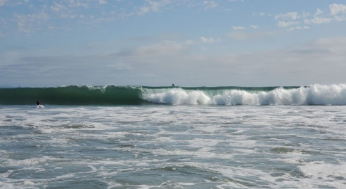 A surfer rides a large wave under a blue sky with scattered clouds, creating a dynamic ocean scene.